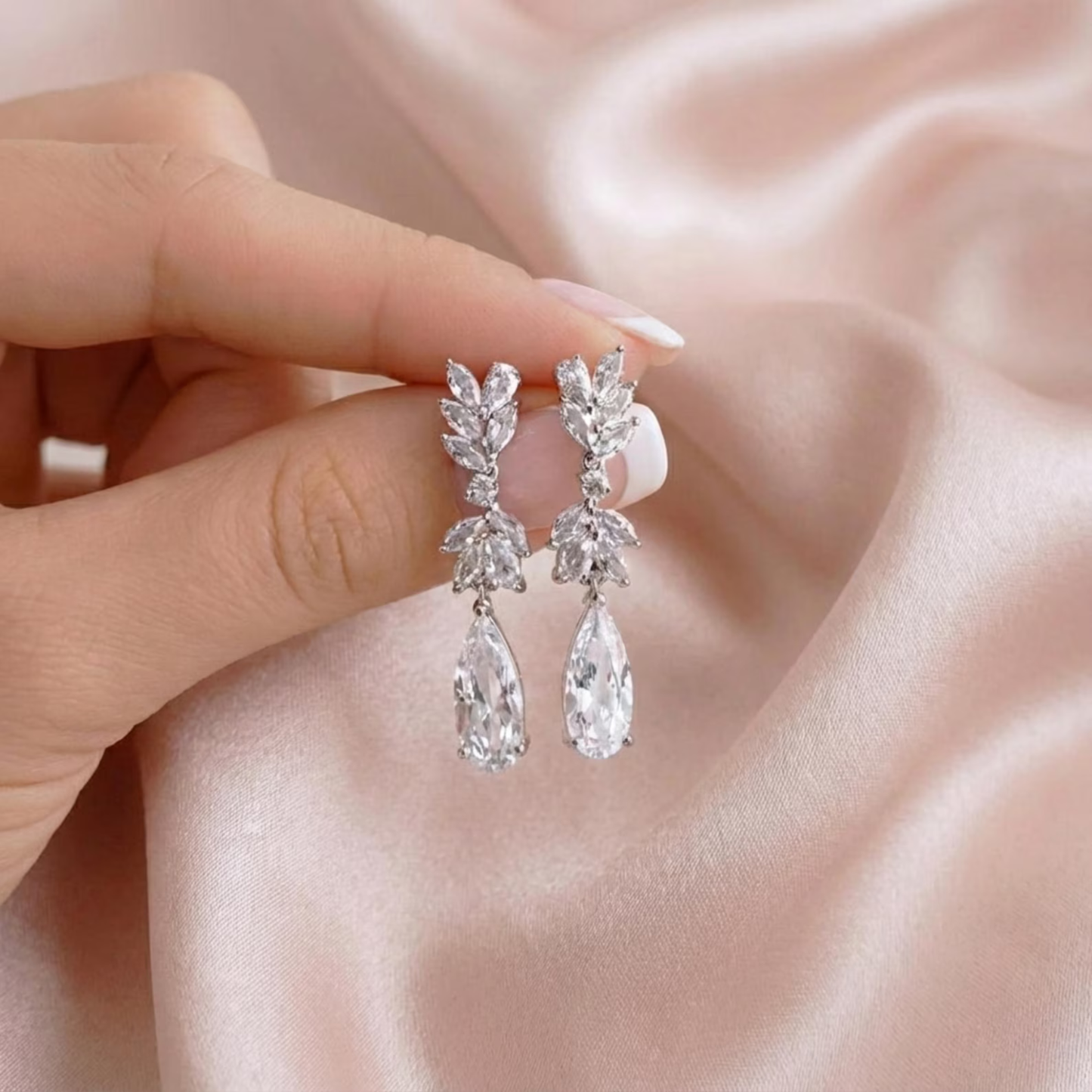 A hand holding a pair of silver cubic zirconia bridal teardrop earrings against a soft pink satin background.