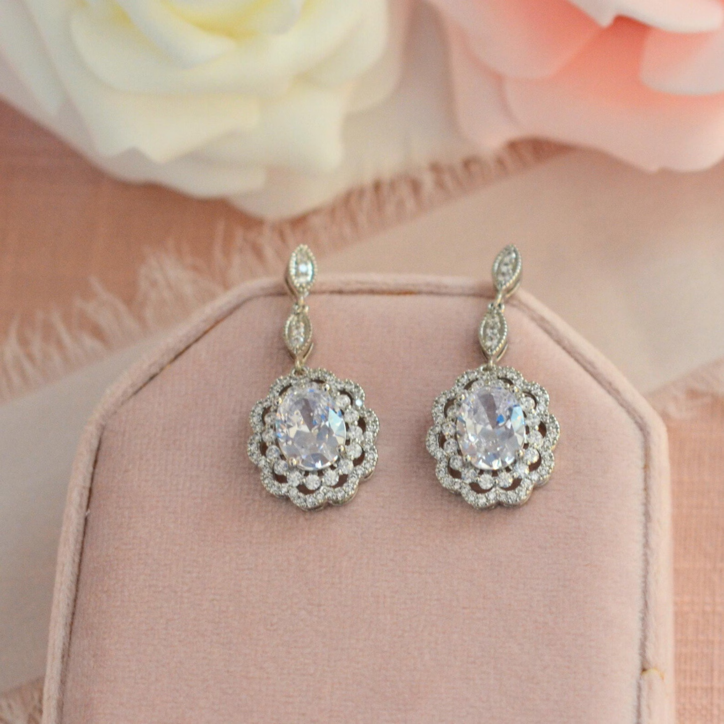 Pair of silver Cubic Zirconia earrings with clear gemstones on a pink cushion with flowers in the background