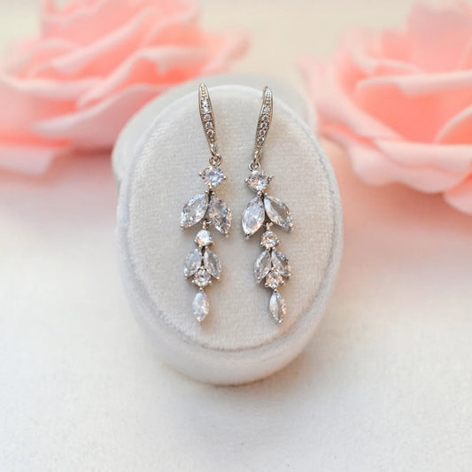 A pair of sterling silver floral drop earrings with sparkling cubic zirconia stones, displayed on a white velvet stand with soft pink roses in the background.